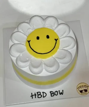 เค้กดอกเดซี่สีเหลืองสดใสพร้อมหน้าที่ยิ้มแย้มและคำว่า HBD BOW สำหรับวันเกิด.
