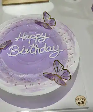 เค้กวันเกิดสีม่วงพร้อมคำว่า Happy Birthday และตกแต่งด้วยผีเสื้อพลาสติกสีม่วงและทองบนเค้ก.