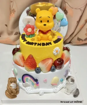 เค้กหมีพูห์ 2 ชั้น สีเหลืองสดใส พร้อมตกแต่งด้วยผลไม้สดและลูกโป่งน่ารัก เหมาะสำหรับงานวันเกิดเด็กและคนรักการ์ตูนหมีพูห์.