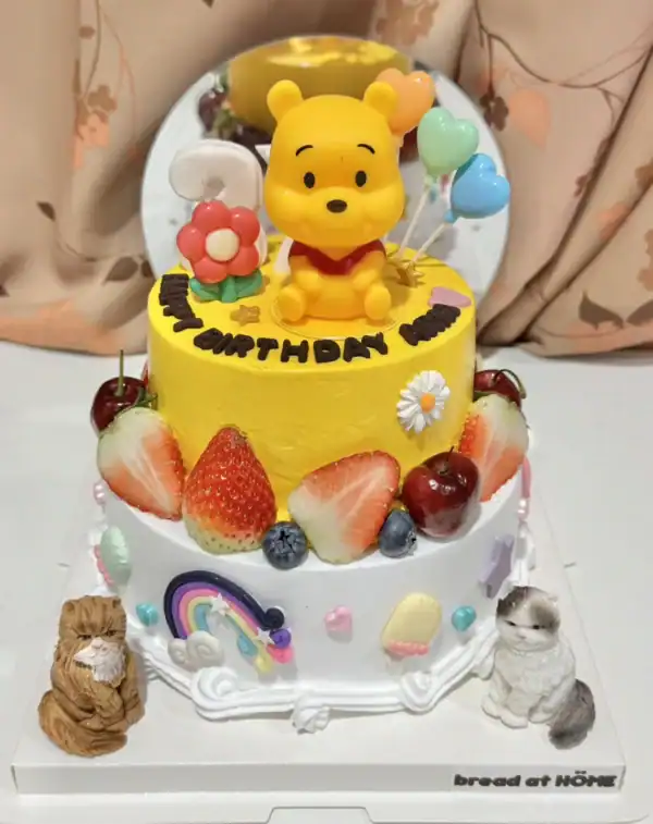 เค้กหมีพูห์ 2 ชั้น สีเหลืองสดใส พร้อมตกแต่งด้วยผลไม้สดและลูกโป่งน่ารัก เหมาะสำหรับงานวันเกิดเด็กและคนรักการ์ตูนหมีพูห์.