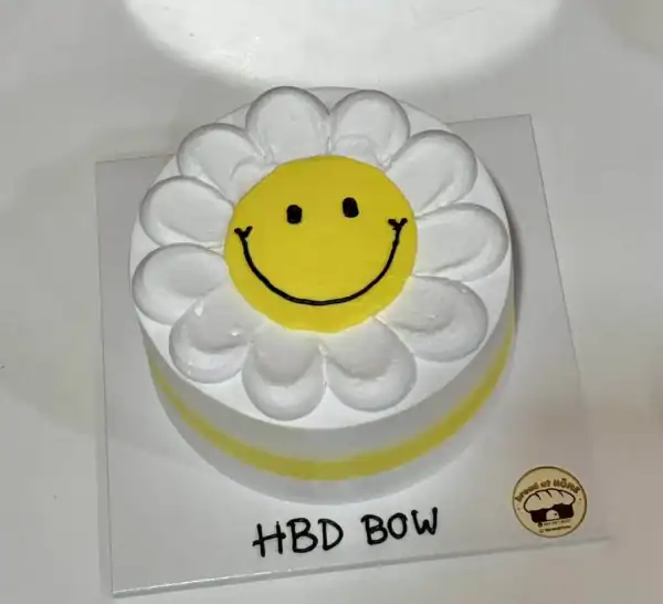 เค้กดอกเดซี่สีเหลืองสดใสพร้อมหน้าที่ยิ้มแย้มและคำว่า HBD BOW สำหรับวันเกิด.