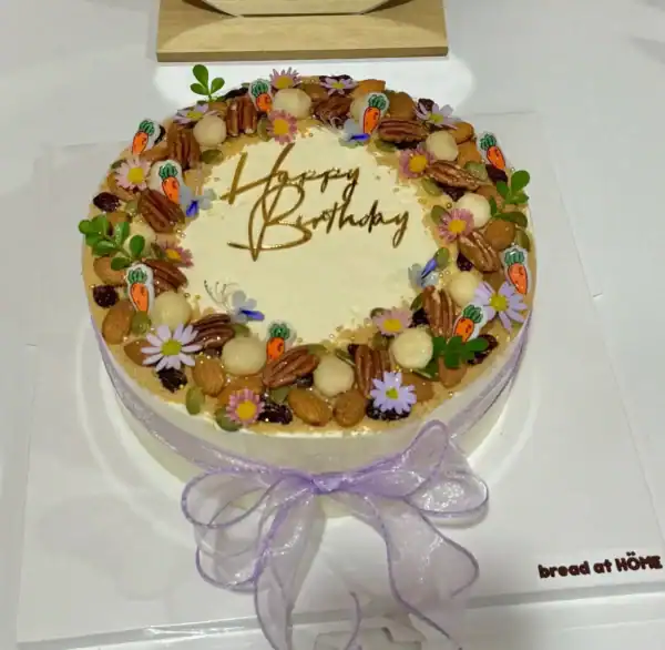 เค้กแครอทเนื้อเนียนนุ่ม พร้อมครีมชีสหวานมัน ตกแต่งด้วยดอกไม้และถั่วหลากสี สวยงามเหมาะสำหรับฉลองวันเกิดหรือโอกาสพิเศษ.