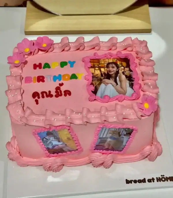 เค้กวันเกิดสีชมพูตกแต่งด้วยดอกไม้และภาพถ่ายของเด็กหญิงในโอกาสพิเศษ พร้อมข้อความสุขสันต์วันเกิดและชื่อ.