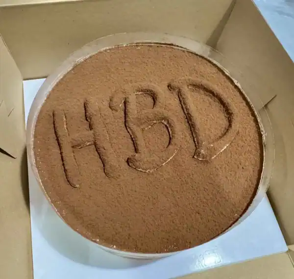 ขนมปังกล่อง HÖME ที่มีคำว่า "HBD" เขียนด้วยผงโกโก้บนหน้าเค้กกลมในกล่องบรรจุภัณฑ์.