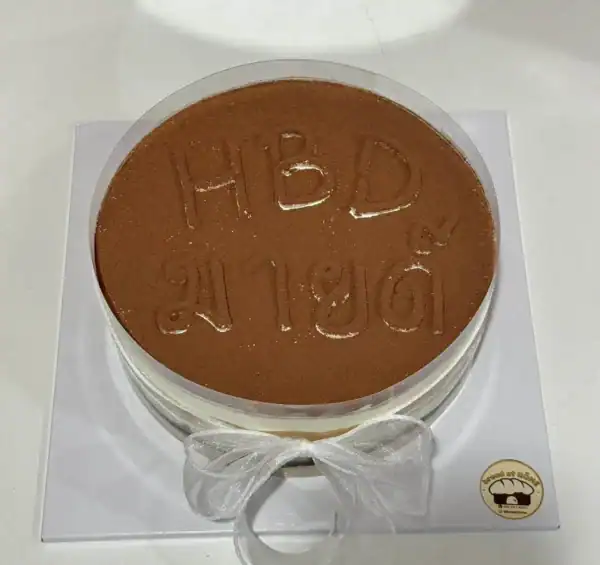 เค้กช็อกโกแลต HBD สำหรับวันเกิด พร้อมตกแต่งด้วยริบบิ้นสีขาวและโลโก้ร้าน Bread at HÖME.