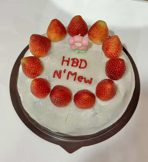 ฟิล์มเนื้อเค้กวันเกิดพร้อมสตรอเบอร์รี่สดสีแดงสดเรียงรอบเค้กและข้อความ "HBD N'Mew" เป็นภาพของขนมเค้กฉลองวันเกิดที่ดูน่ารับประทานในบรรยากาศบ้านในประเทศไทย.