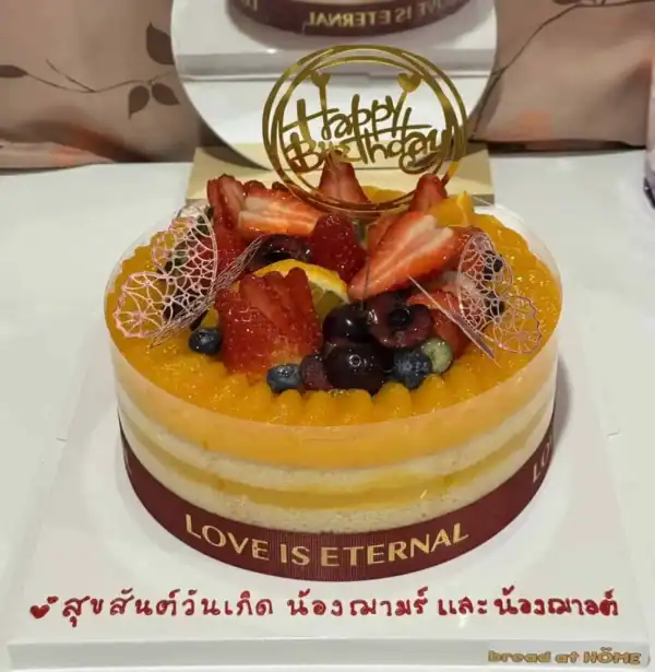 เค้กส้มหน้านิ่มพรีเมียมตกแต่งด้วยผลไม้สดและลายลูกไม้บนเค้ก พร้อมคำว่า Happy Birthday บนยอดเค้ก.