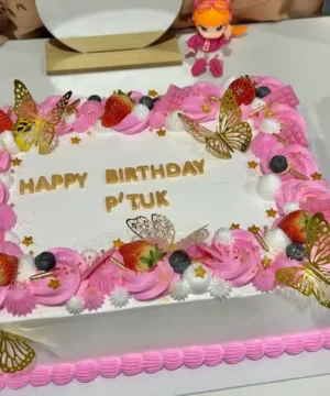 เค้กวันเกิดสีชมพูสดใส ตกแต่งด้วยครีมและผลไม้สด เช่น สตรอเบอร์รี่และบลูเบอร์รี่ พร้อมดีไซน์ผีเสื้อทองคำและดอกไม้เล็กๆ ให้ความรู้สึกน่ารักและอร่อย.
