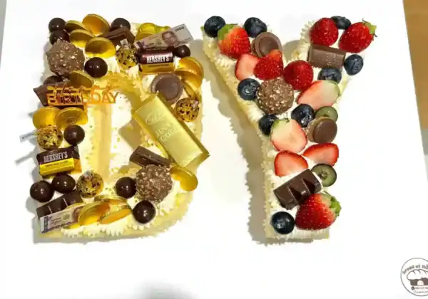 เค้กตัวอักษรวันเกิดทำจากครีมและผลไม้สด พร้อมตกแต่งด้วยช็อกโกแลตและขนมหวานหลากสี เหมาะสำหรับงานฉลองวันเกิดที่มีความหมายดีและดีไซน์เก๋ไก๋.