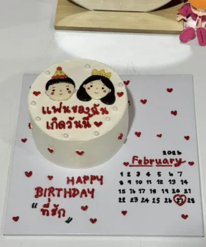 เค้กวันเกิดคู่รักพร้อมภาพการ์ตูนเจ้าบ่าวเจ้าสาวและข้อความในภาษาไทย พร้อมตกแต่งด้วยหัวใจสีแดงบนเค้กและฐานรองเค้ก.