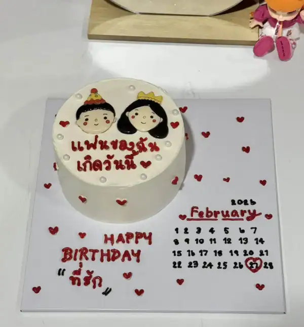เค้กวันเกิดคู่รักพร้อมภาพการ์ตูนเจ้าบ่าวเจ้าสาวและข้อความในภาษาไทย พร้อมตกแต่งด้วยหัวใจสีแดงบนเค้กและฐานรองเค้ก.