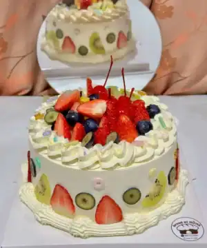 เค้กผลไม้สดรวมที่เต็มไปด้วยผลไม้หลากหลาย เช่น สตรอเบอร์รี่ กีวี บลูเบอร์รี่ และเชอร์รี่ บนเค้กเนื้อเนียนนุ่มตกแต่งด้วยครีมและลูกกวาดสีสันสดใส.