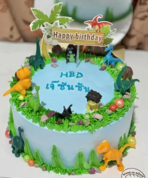 ก้อนเค้กวันเกิดโปรโมตความสนุกสนานด้วยธีมไดโนเสาร์สีสันสดใสและเทคนิคการตกแต่งด้วยครีมและของตกแต่งน่ารักบนแผ่นเค้กสีฟ้า.