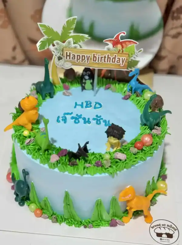 ก้อนเค้กวันเกิดโปรโมตความสนุกสนานด้วยธีมไดโนเสาร์สีสันสดใสและเทคนิคการตกแต่งด้วยครีมและของตกแต่งน่ารักบนแผ่นเค้กสีฟ้า.