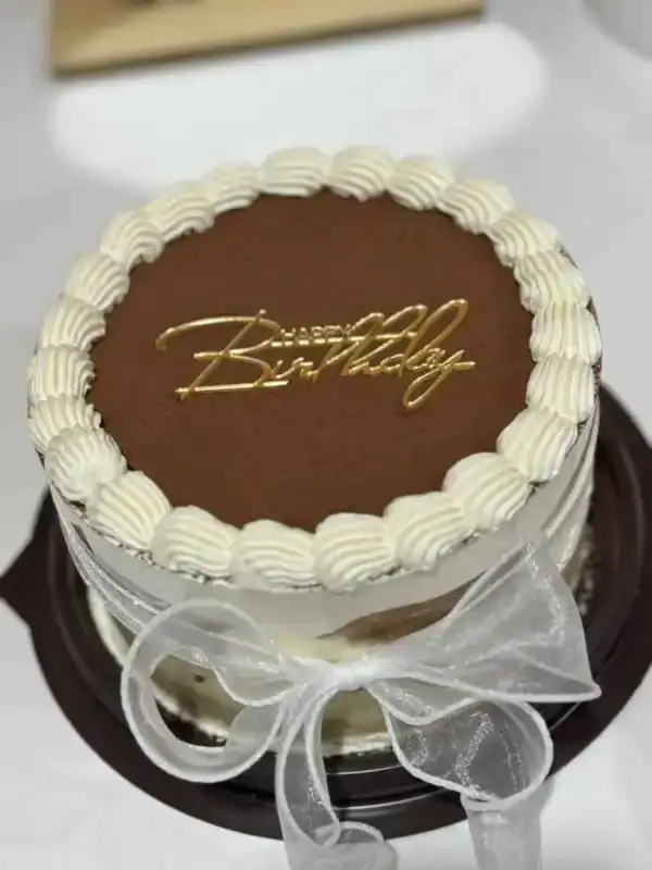 เค้กทีรามิสุเนื้อเนียนนุ่ม ปกคลุมด้วยช็อกโกแลตและตกแต่งด้วยคำว่า 'Birthday' ด้วยสีทอง สวยงามและน่ากิน.