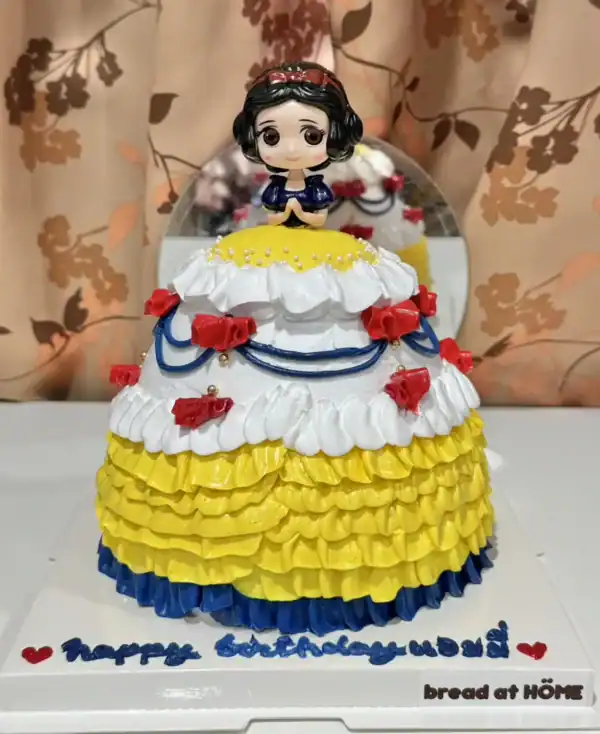 เค้กสโนไวท์หวานละมุนตกแต่งด้วยครีมและลูกกวาดสีสดใส พร้อมตัวตุ๊กตาน่ารักบนยอดเค้ก เหมาะสำหรับงานฉลองและวันเกิดที่ต้องการความน่ารักและความหวาน.